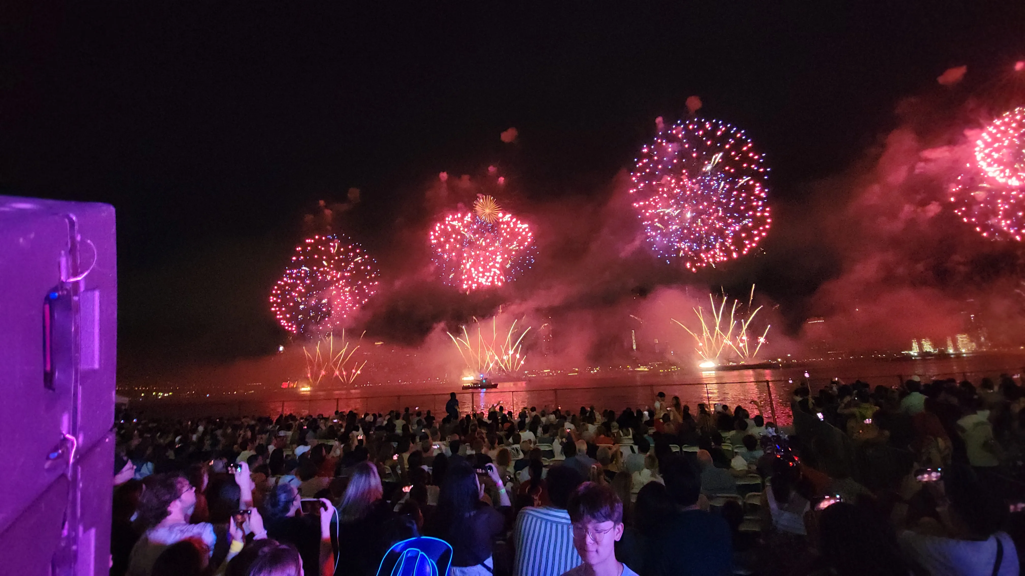 Red fireworks bursts over water with large seated crowd